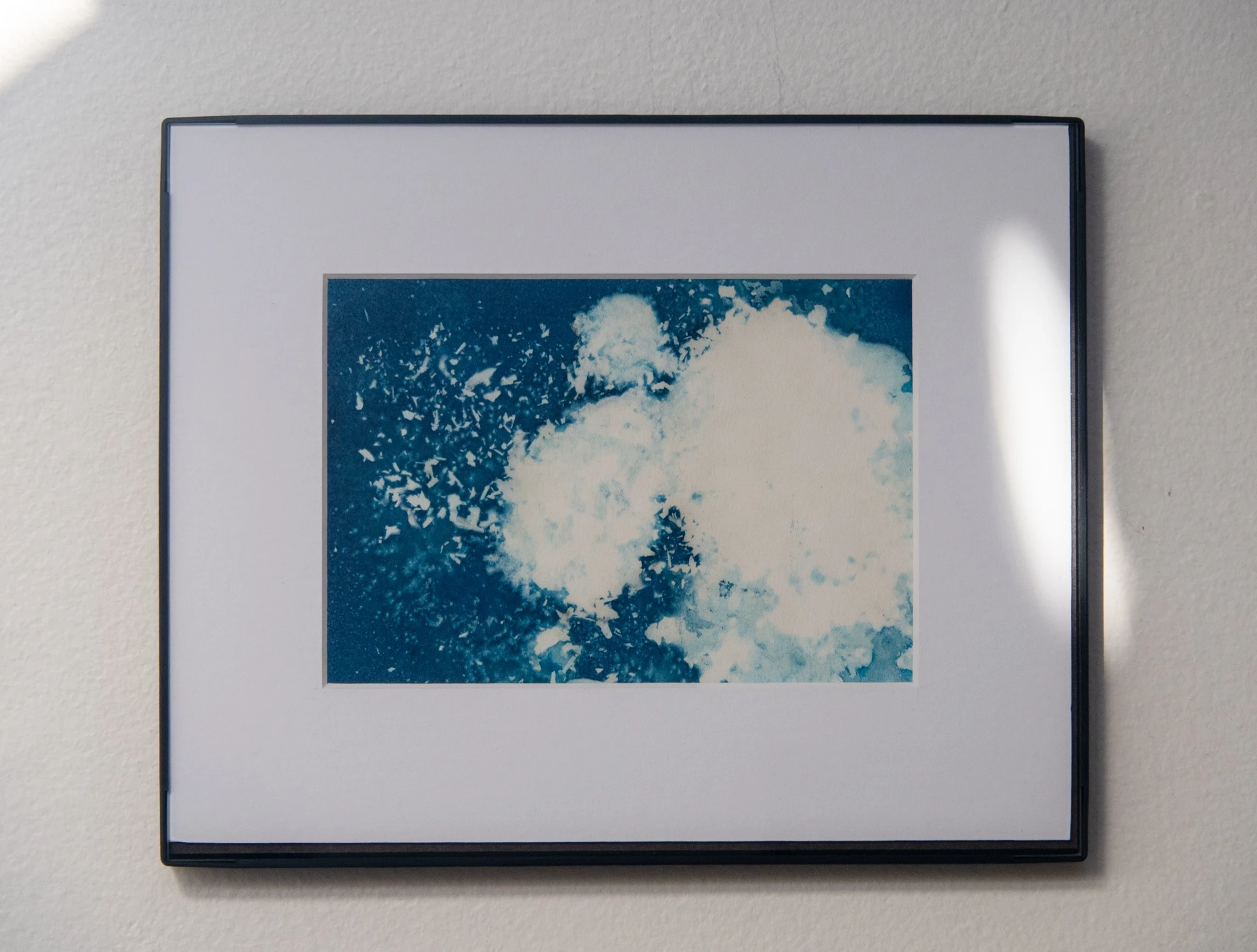 The print framed in a slim black frame with a wide white mat, hanging on a pale wall in raked afternoon light.
